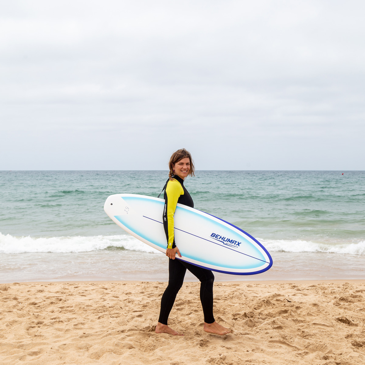 Una mujer con traje de neopreno camina por una playa de arena cargando una tabla de surf, con el mar al fondo.