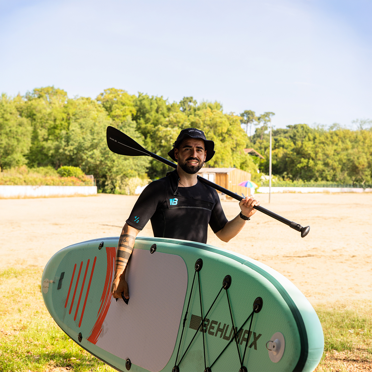 Un hombre con traje de neopreno sostiene una tabla de SUP verde menta y un remo en la playa.
