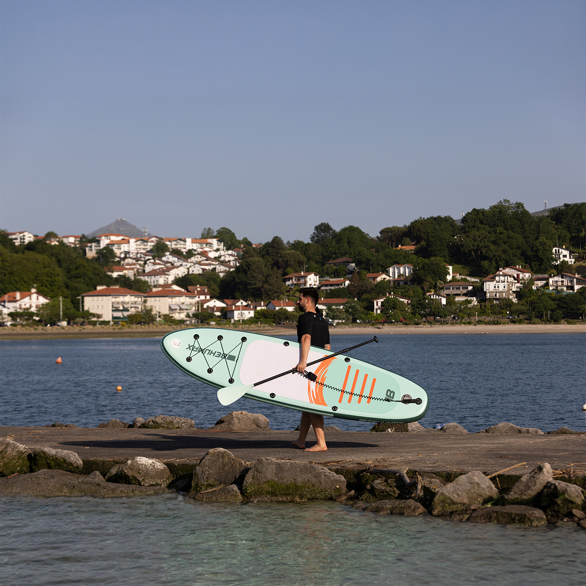 Un hombre lleva una tabla de paddle surf turquesa por la orilla.