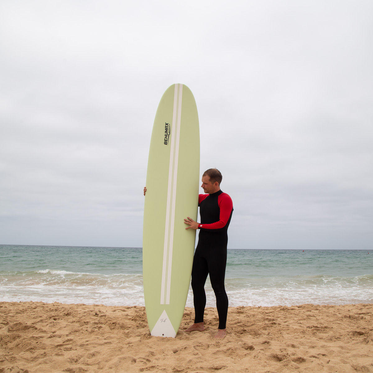 Un surfista con traje negro y mangas rojas sostiene una tabla de surf alta y verde claro en una playa de arena.