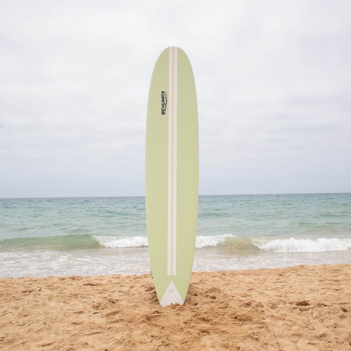 Una tabla de surf verde claro se encuentra erguida en la arena de una playa con el mar y cielo nublado de fondo.