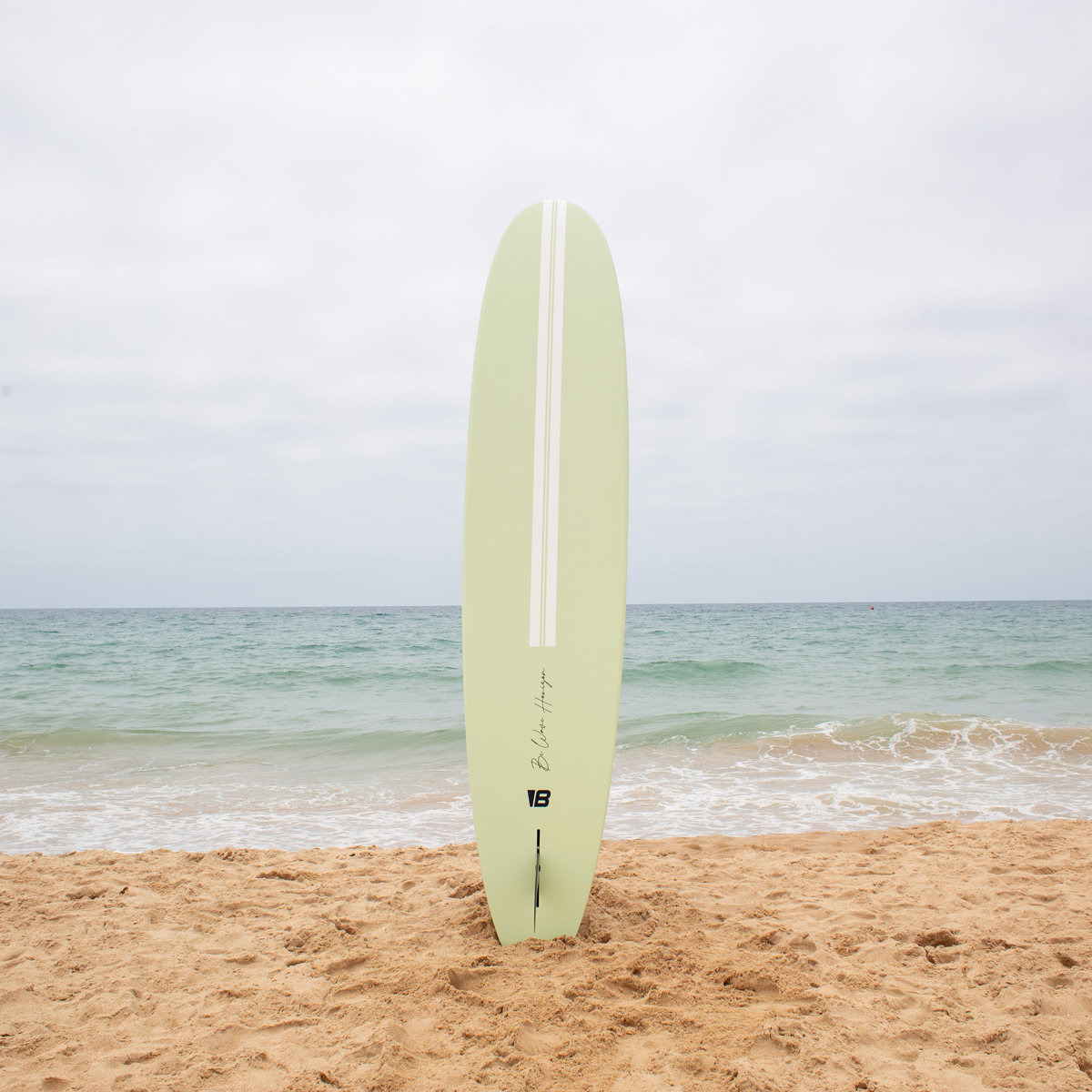 Una tabla de surf verde claro se encuentra verticalmente en la arena de una playa ante el mar bajo un cielo nublado.