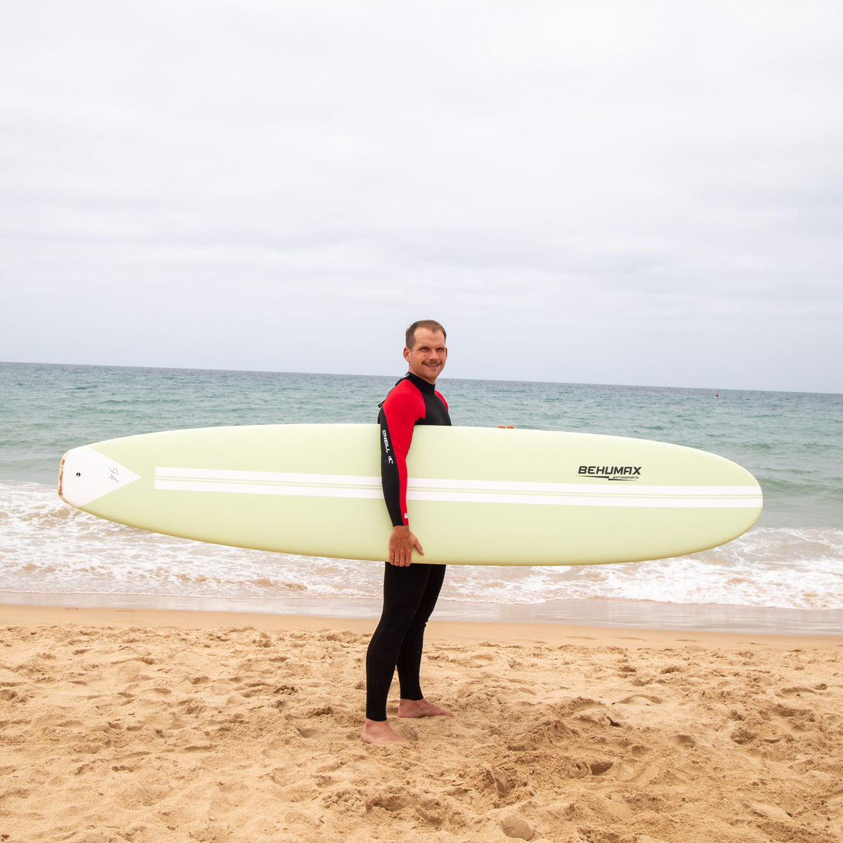 Un surfista con traje de neopreno sostiene una tabla verde clara en una playa de arena ante el mar.