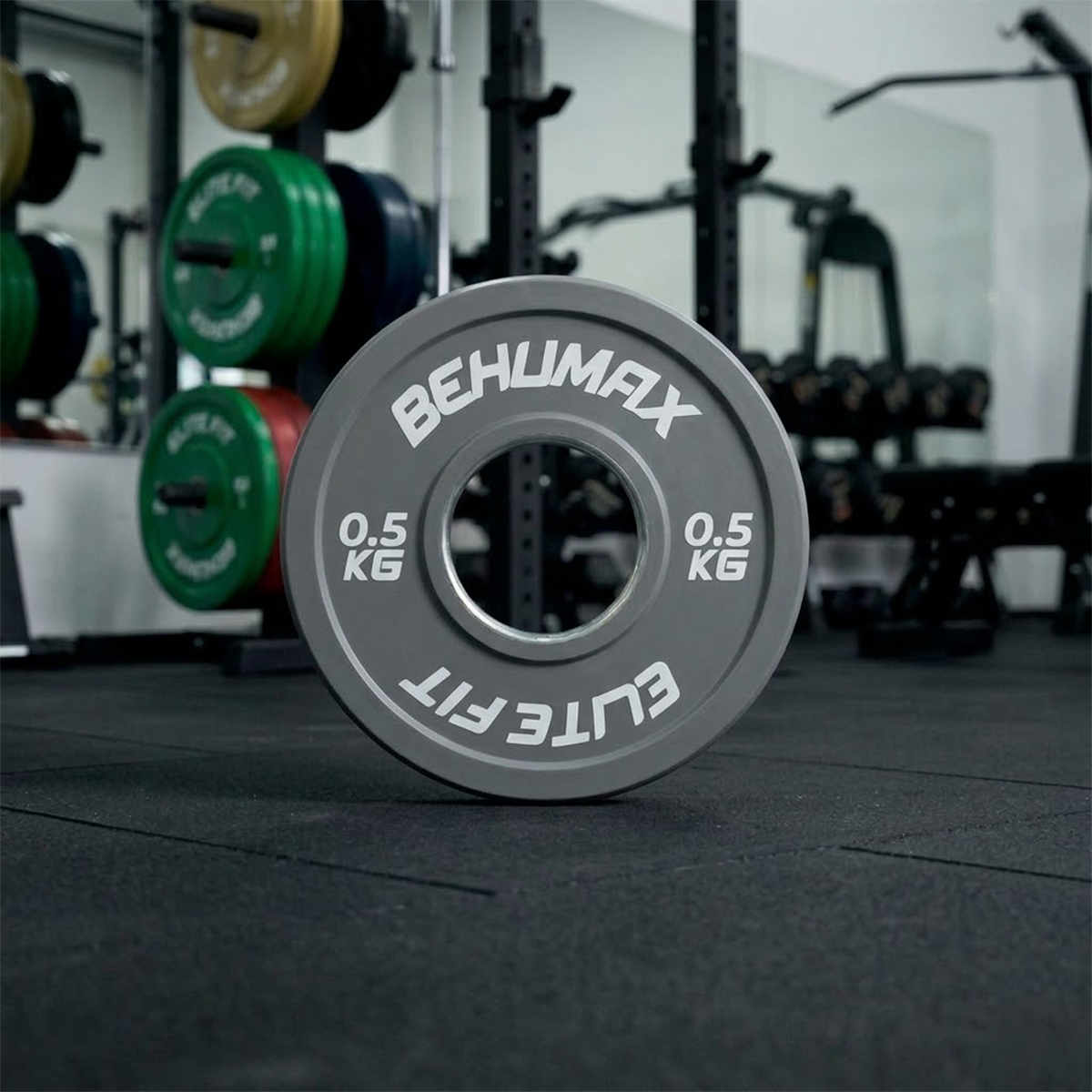 Un disco de pesas gris de 0.5 kg con texto descansa en el suelo del gimnasio, con más pesas al fondo.
