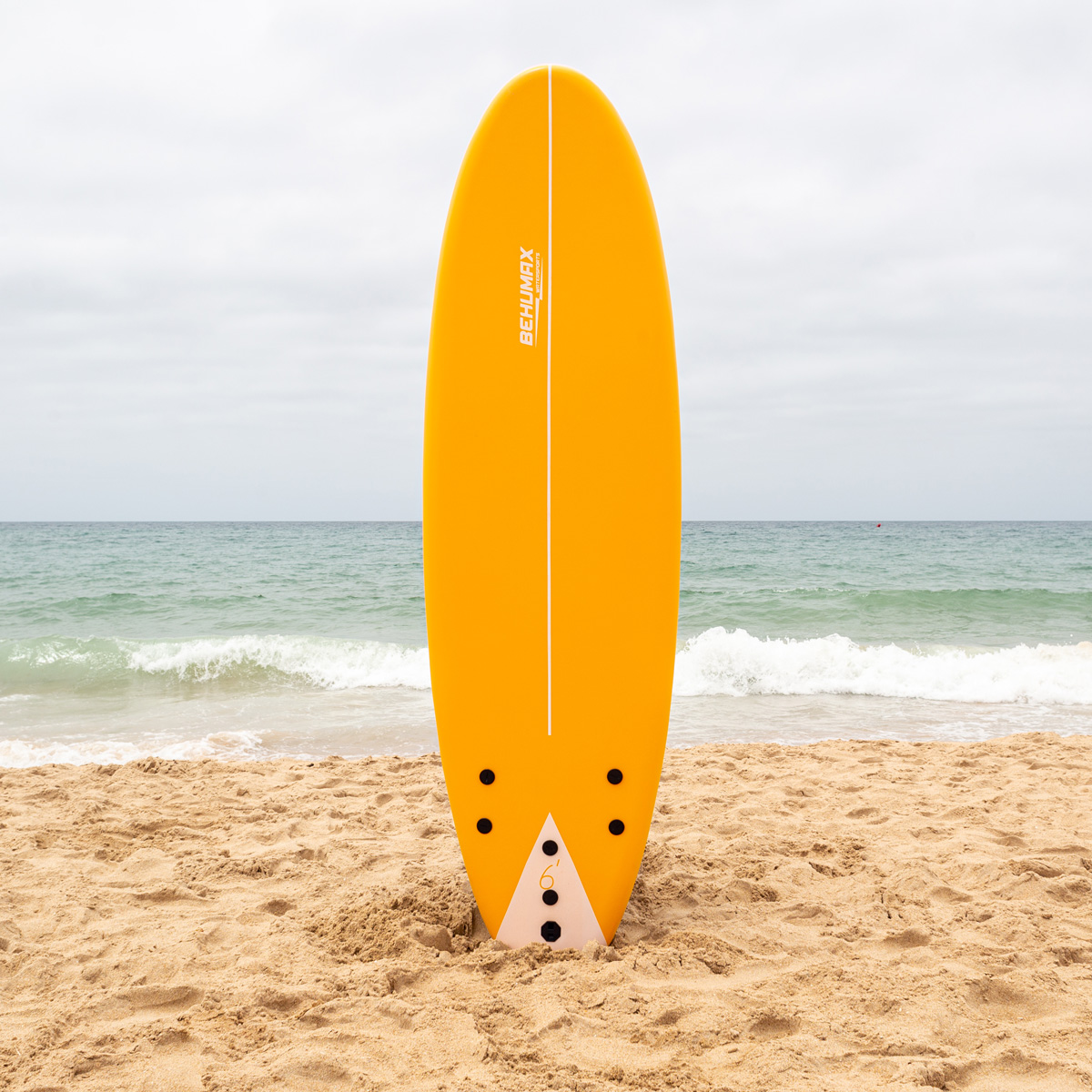 Una tabla de surf amarilla se encuentra verticalmente en la arena de una playa con el mar tranquilo.