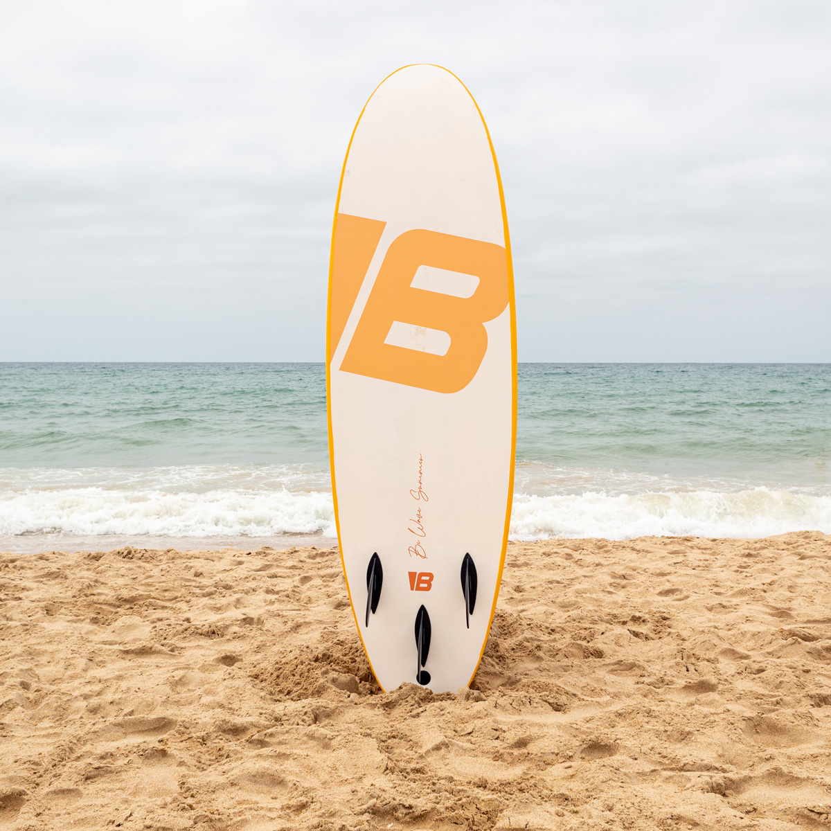 Una tabla de surf blanca y amarilla con una gran letra B está vertical en la arena de una playa frente al mar.