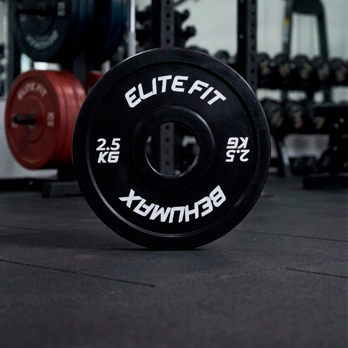 Un disco de pesas negro de 2,5 kg con marca Elite Fit en el suelo de un gimnasio, con otros pesos desenfocados.