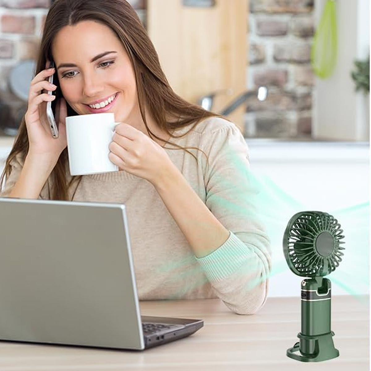Frau telefoniert und trinkt Kaffee vor einem Laptop, neben einem grünen Tischventilator auf dem Schreibtisch.
