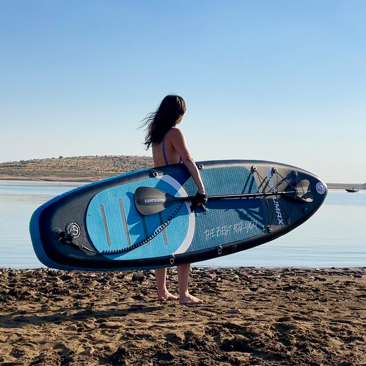 Una mujer sostiene una tabla de paddle surf azul en una playa de arena junto a un lago tranquilo.
