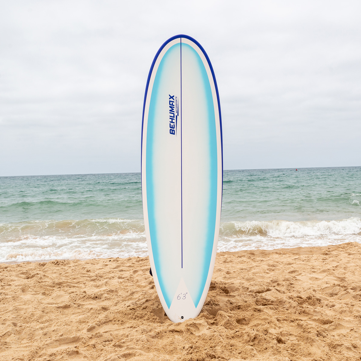 Una tabla de surf blanca y azul se alza verticalmente en una playa de arena frente al mar y cielo nublado.