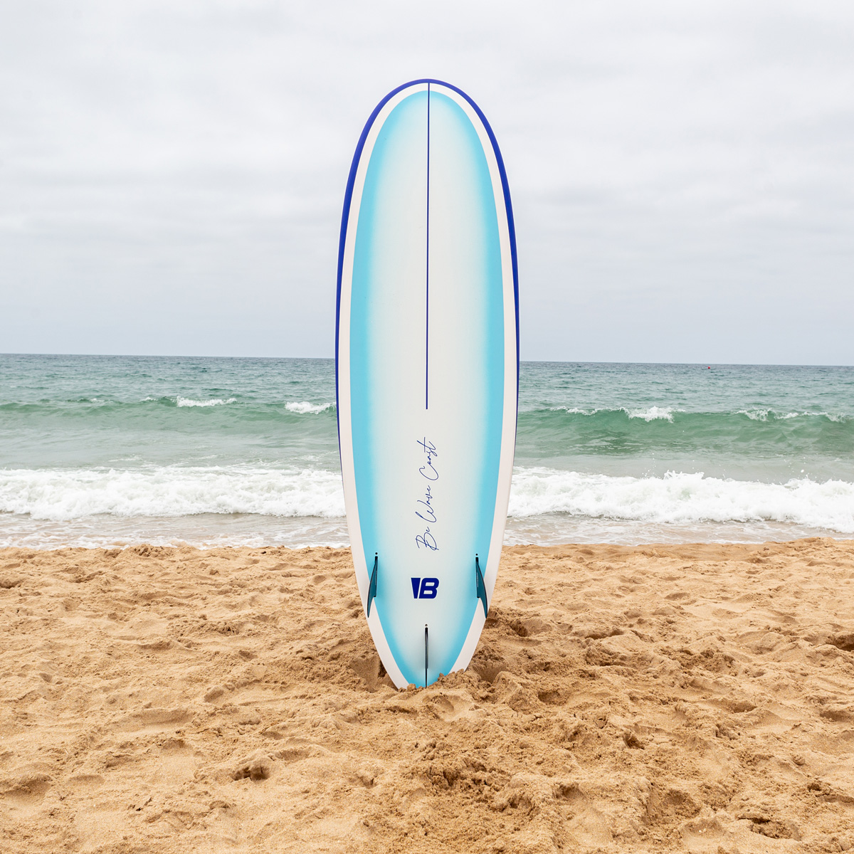Una tabla de surf azul y blanca clavada en la arena de una playa con olas del mar al fondo.