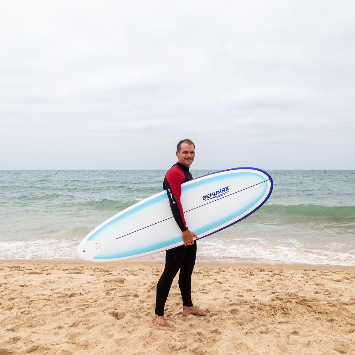Un hombre con traje de neopreno está en una playa de arena sosteniendo una tabla de surf horizontalmente.