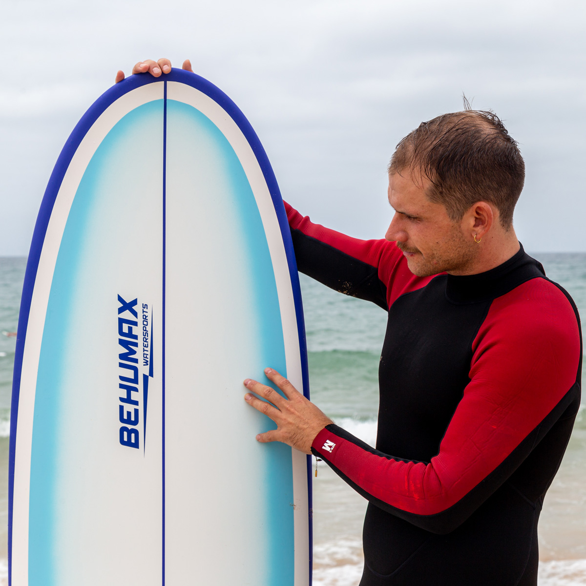 Un hombre con traje de neopreno negro sostiene una tabla de surf blanca y azul en una playa.