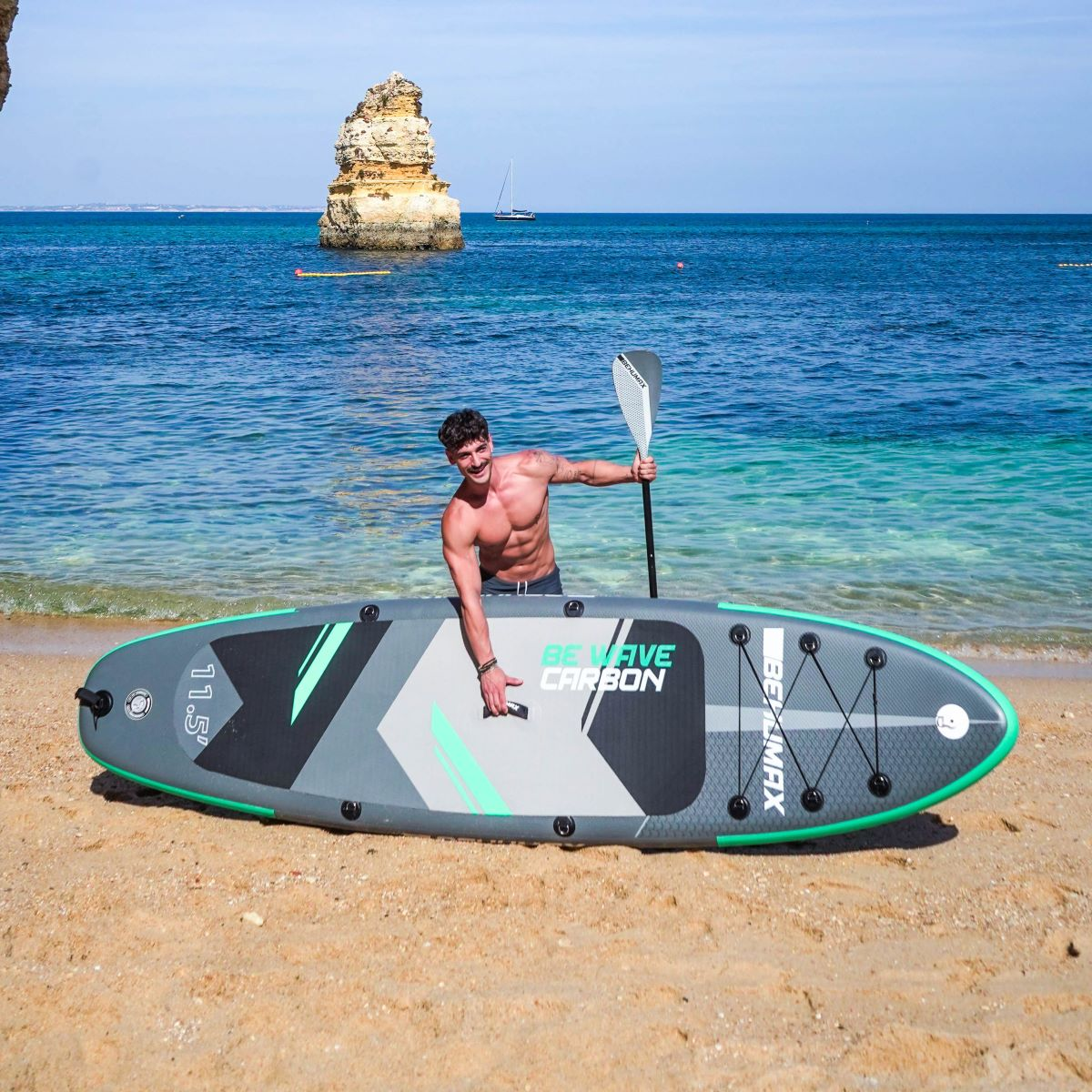Un hombre posa en la playa con una tabla de surf de remo, con el mar azul y un islote rocoso al fondo.