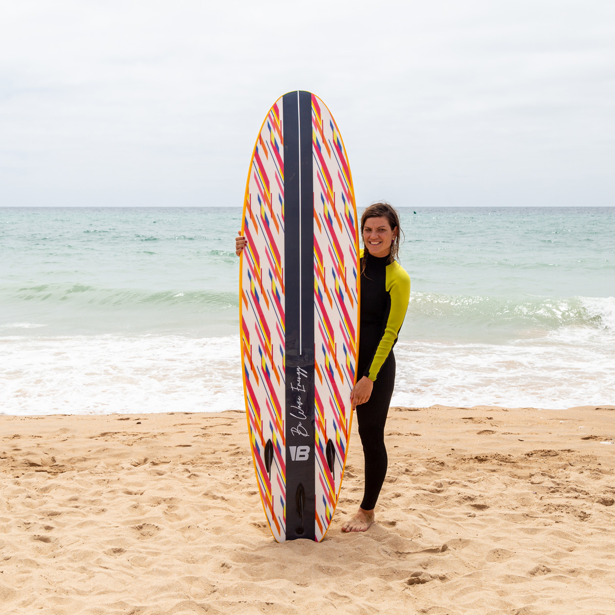 Una mujer con traje de neopreno sostiene una tabla de surf colorida en una playa de arena con olas.