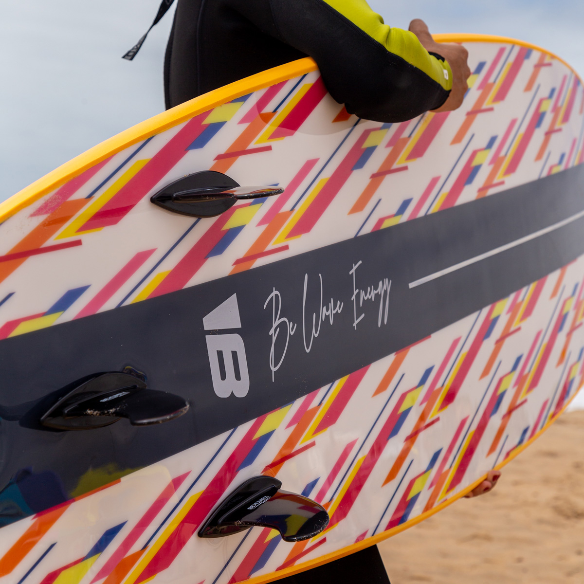 Una persona con traje de neopreno sostiene una tabla de surf colorida con patrones geométricos y tres aletas negras.