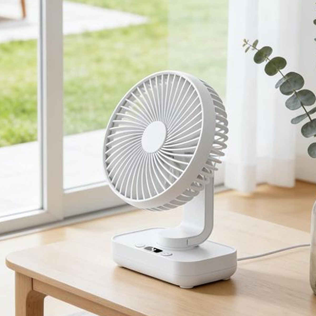 Ein kleiner weißer Tischventilator steht auf einem Holzbrett vor einem Fenster.