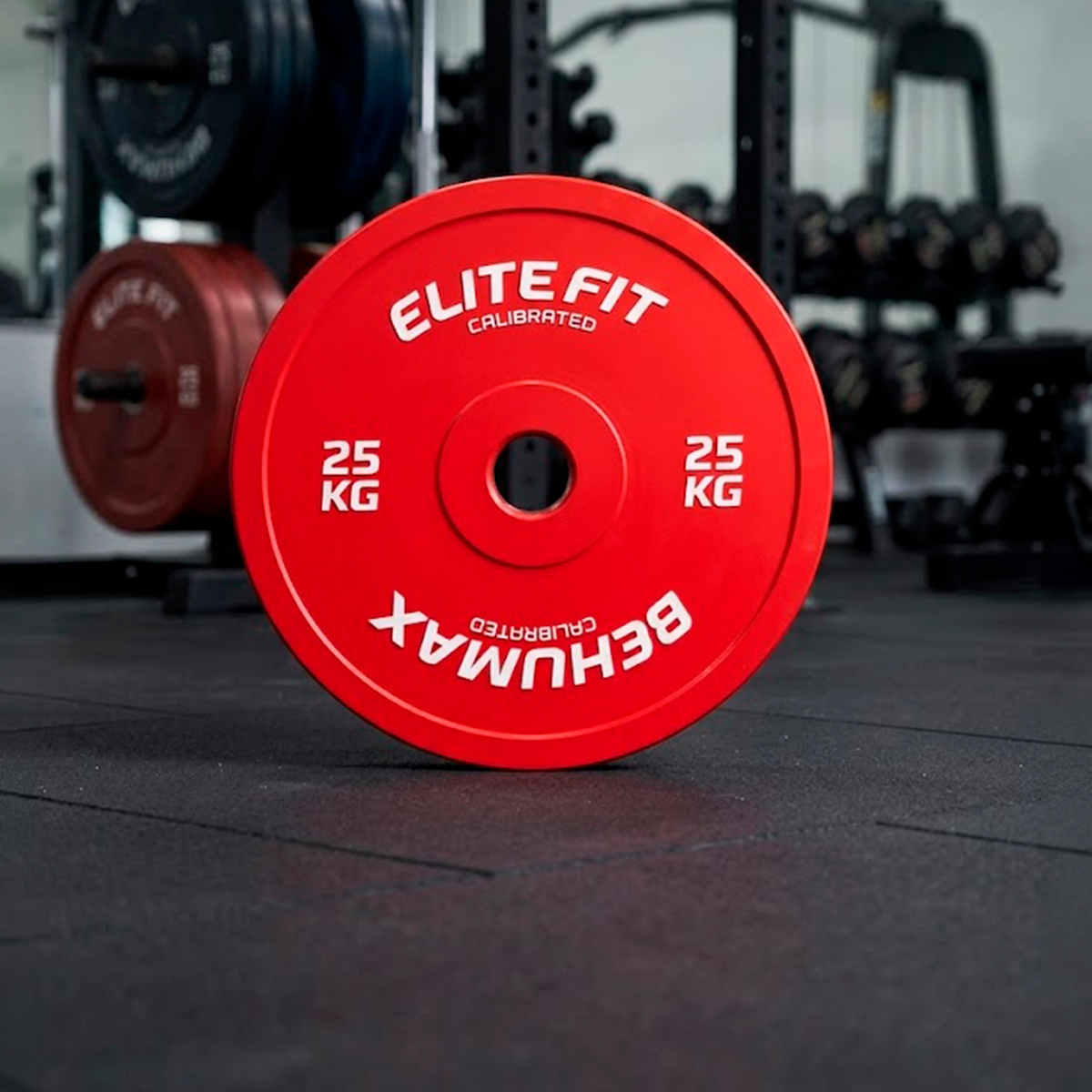 Un disco de peso rojo calibrado Elitefit de 25 kg vertical en el suelo del gimnasio, con pesas y soportes al fondo.