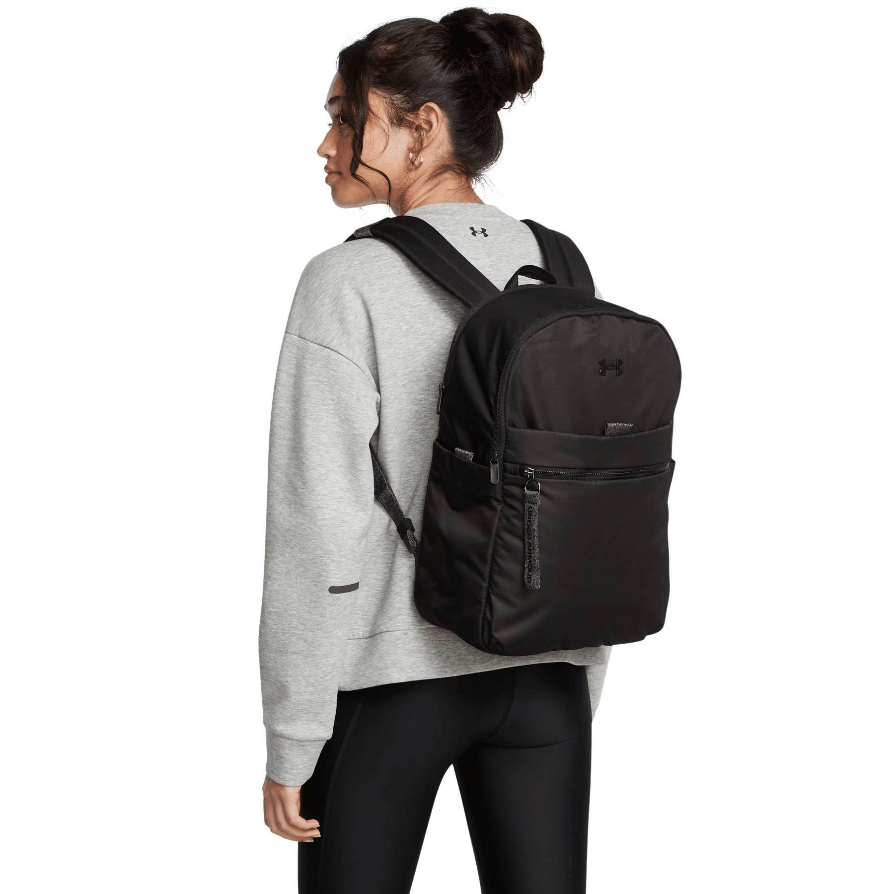 Eine Frau in grauem Pullover und schwarzer Leggings trägt einen schwarzen Under Armour Rucksack vor schwarzem Hintergrund.
