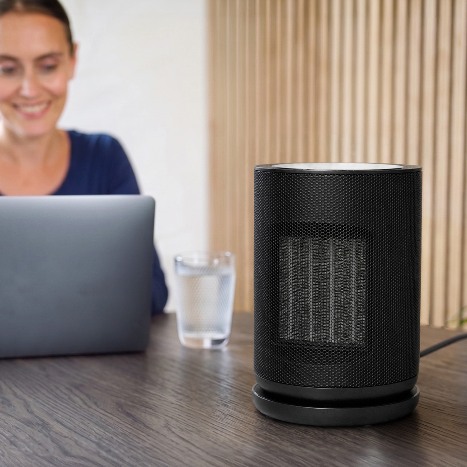 Ein schwarzer Heizlüfter steht auf einem Holztisch, im Hintergrund eine Frau am Laptop und ein Glas Wasser.