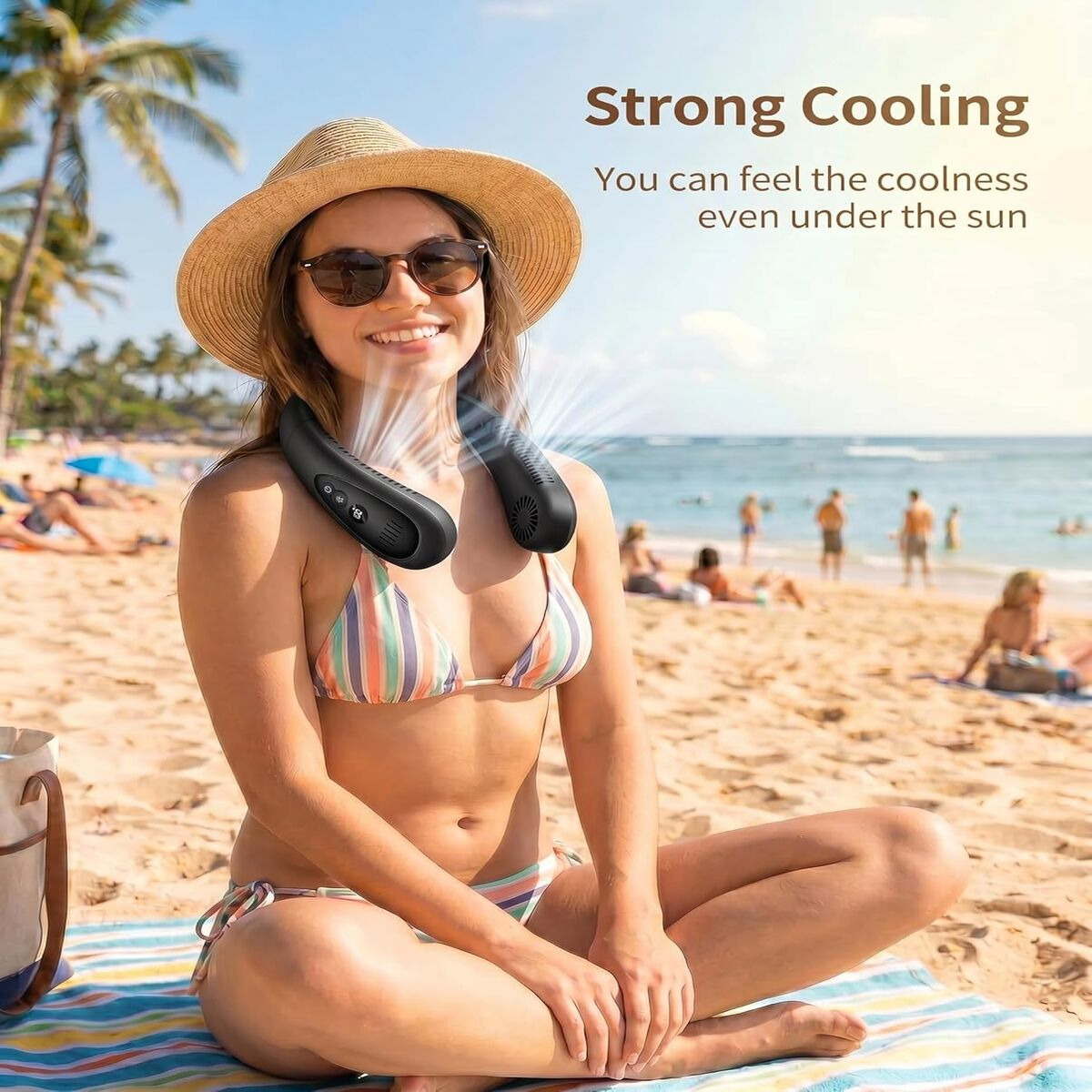 Eine Frau im Bikini mit Strohhut und Sonnenbrille sitzt am Strand und trägt einen schwarzen Nackenventilator.