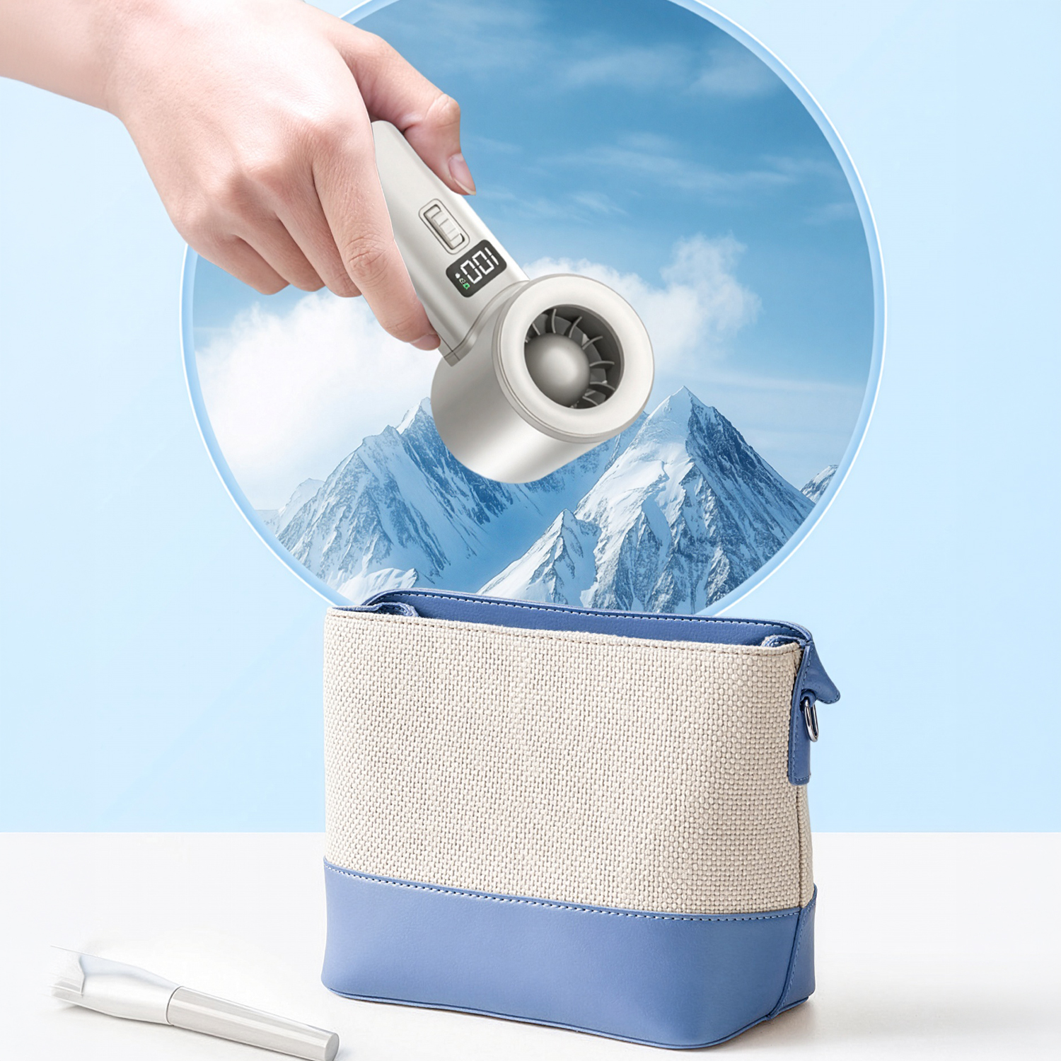 Una mano sostiene un ventilador portátil blanco sobre un neceser azul y beige frente a montañas nevadas.