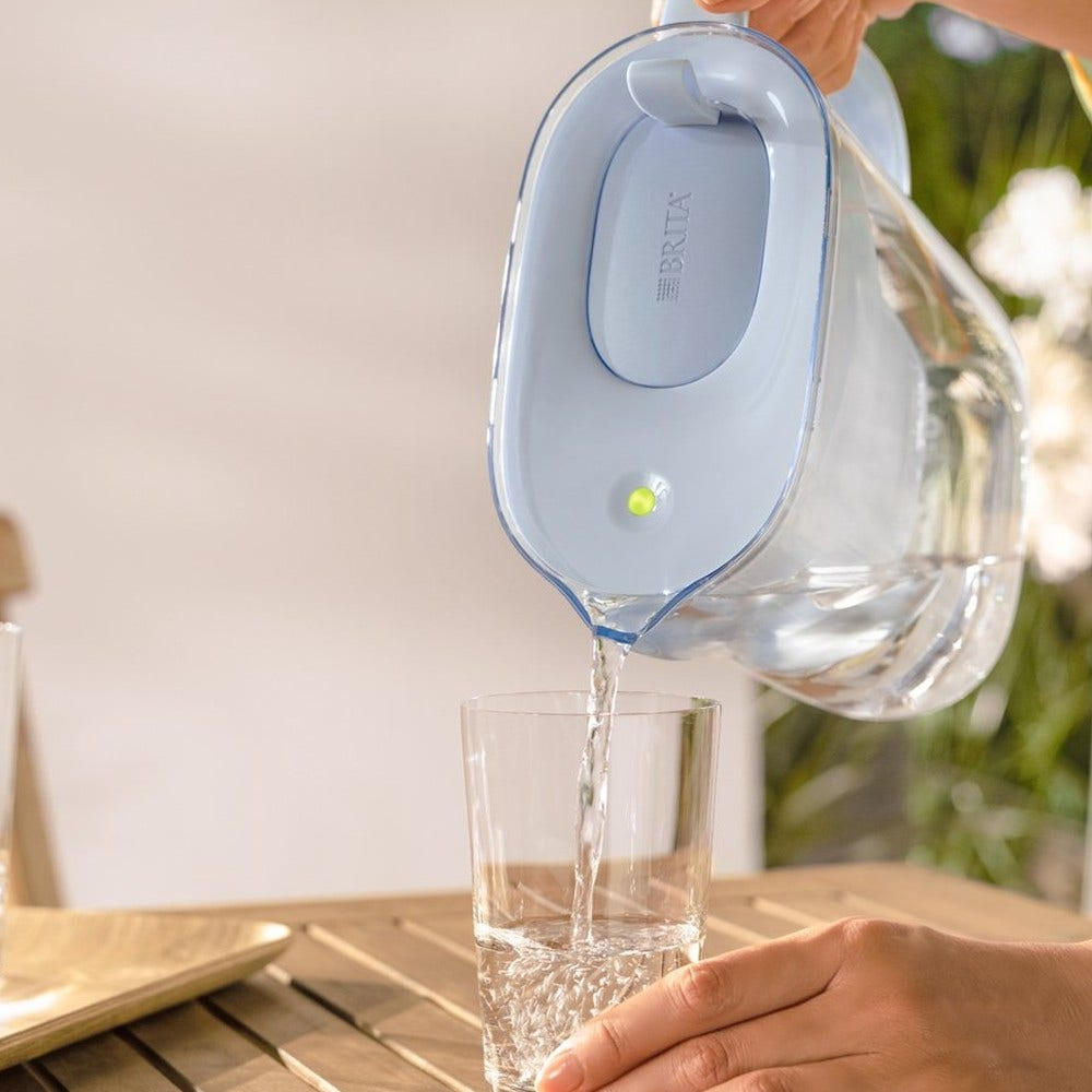 Una mano versa acqua da una caraffa Brita azzurra in un bicchiere su un tavolo di legno.