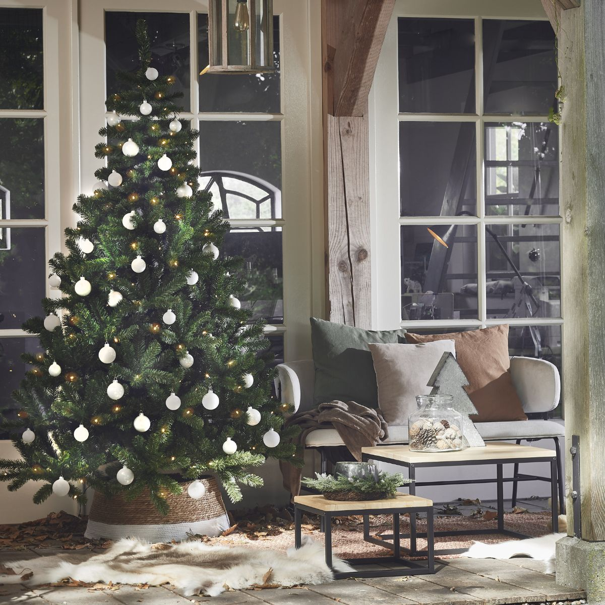 Verlichte kerstboom met witte kerstballen in een gezellige kamer met bank en tafels.