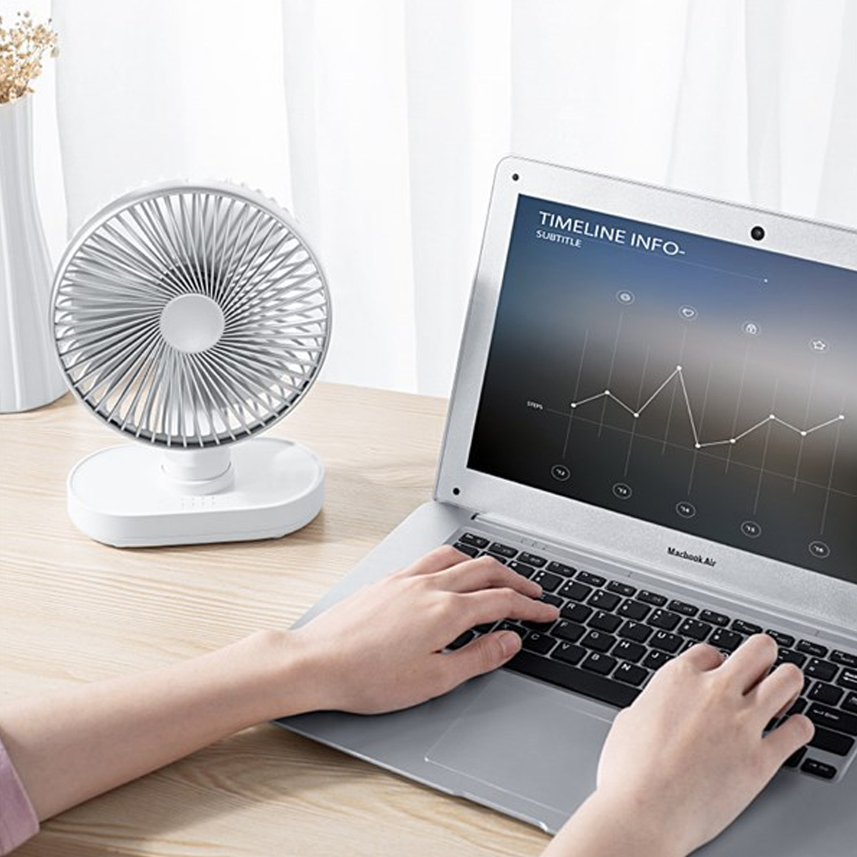 Ein kleiner weißer Tischventilator steht neben einem Laptop mit Händen auf der Tastatur auf einem Holztisch.