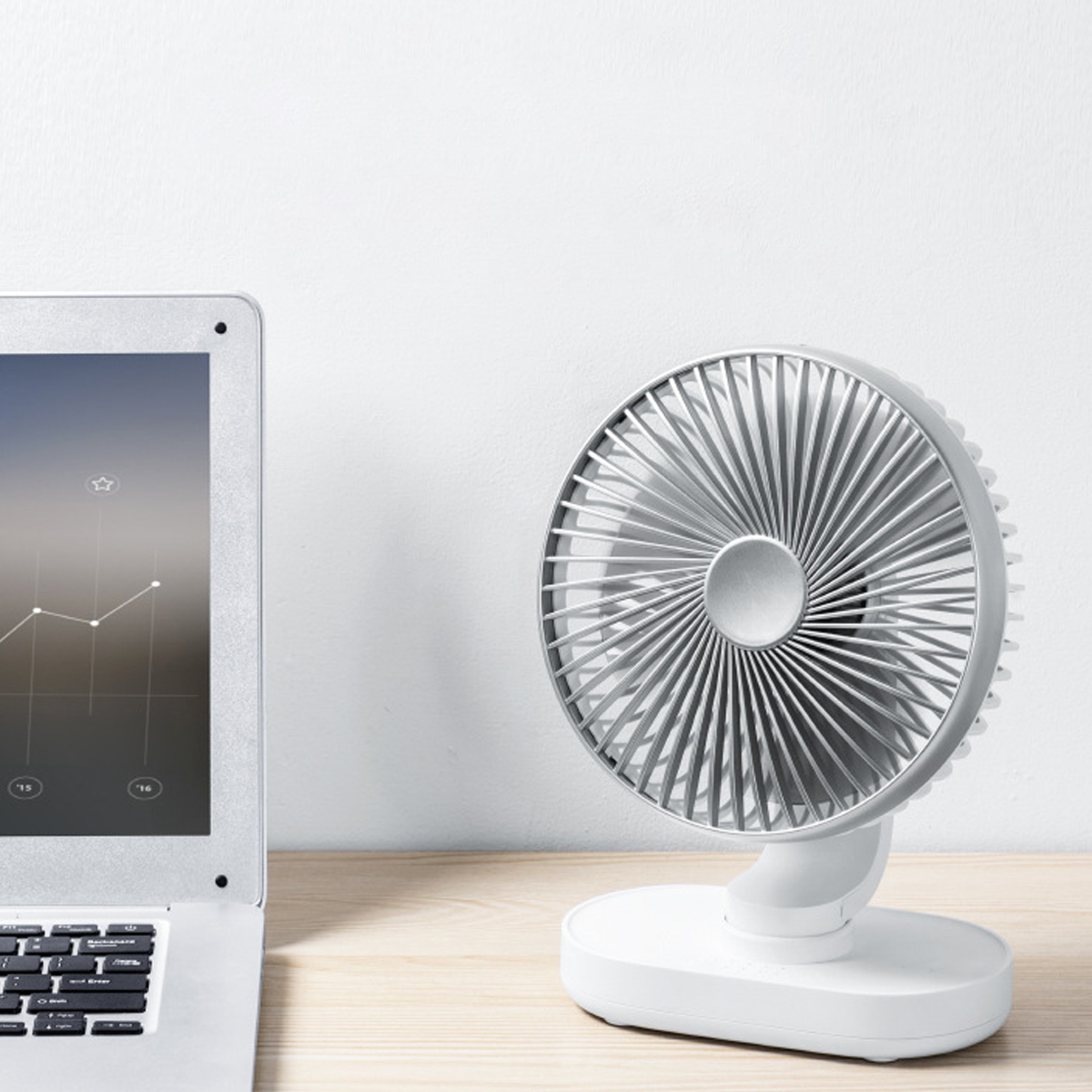 Ein kleiner silberner Tischventilator und ein Laptop auf einem Holztisch vor einer weißen Wand.