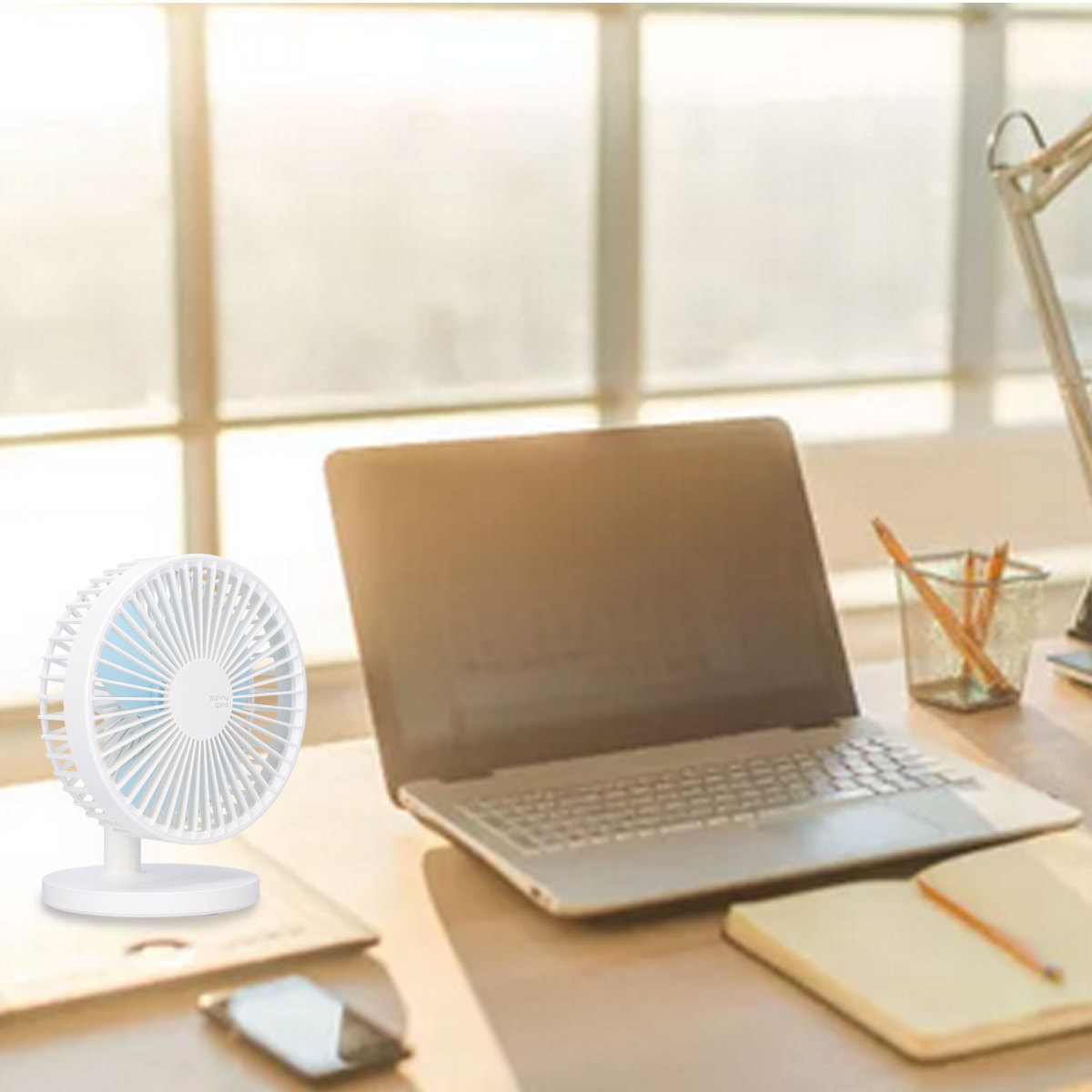 Ein kleiner weißer Schreibtischventilator neben einem offenen Laptop auf einem sonnigen Schreibtisch.