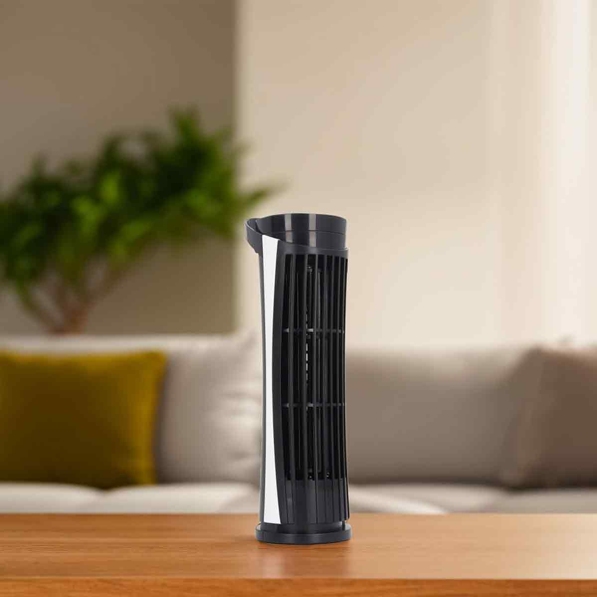 Ein schwarzer Turmventilator steht auf einem Holztisch in einem hellen Wohnzimmer mit Sofa und grüner Pflanze im Hintergrund.