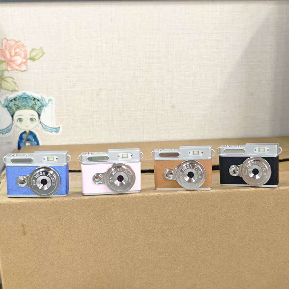 Vier kleine Spielzeugkameras in Blau, Rosa, Braun und Schwarz stehen nebeneinander auf einer Pappschachtel.