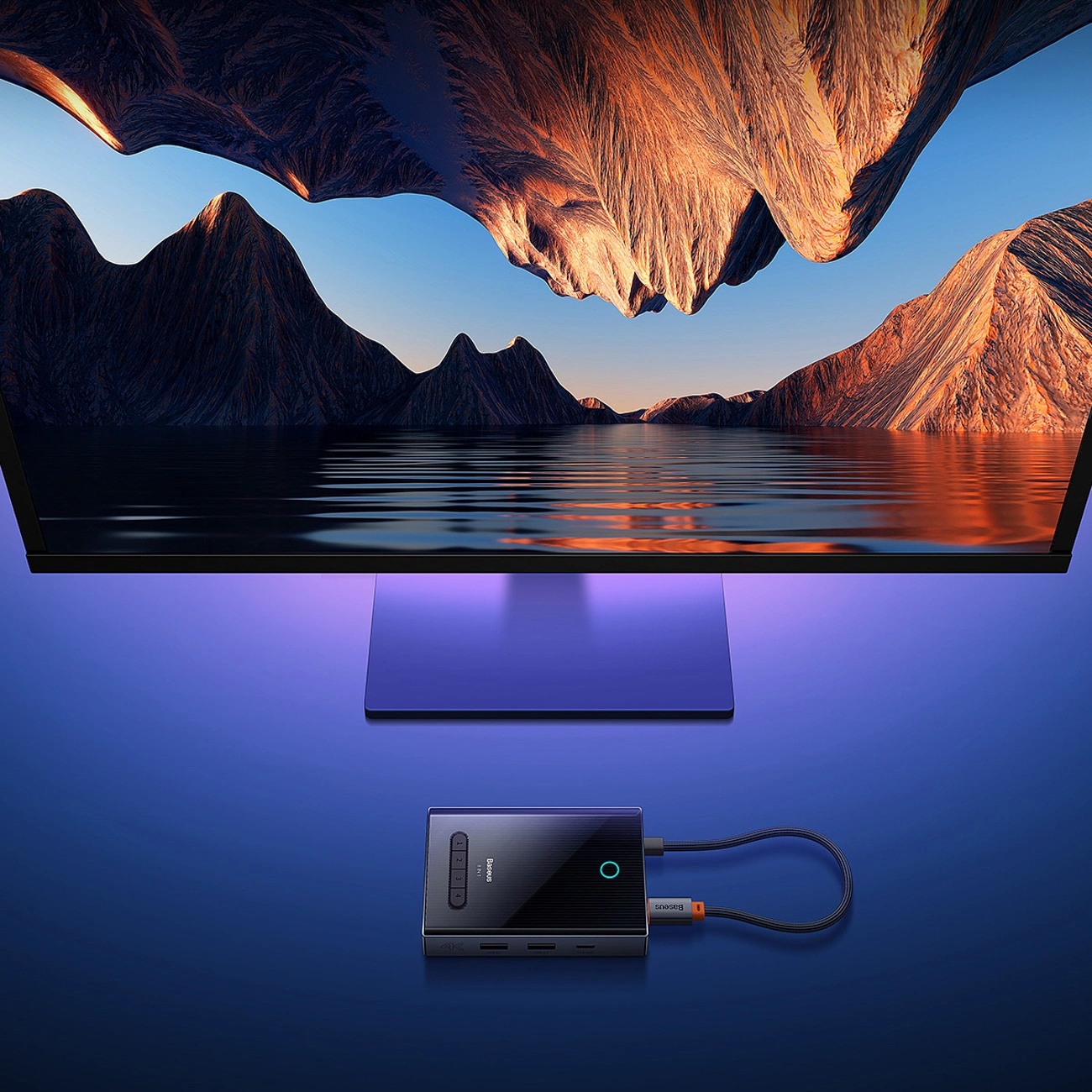 Un hub USB-C grigio si trova su una scrivania blu davanti a un monitor con paesaggio montano.