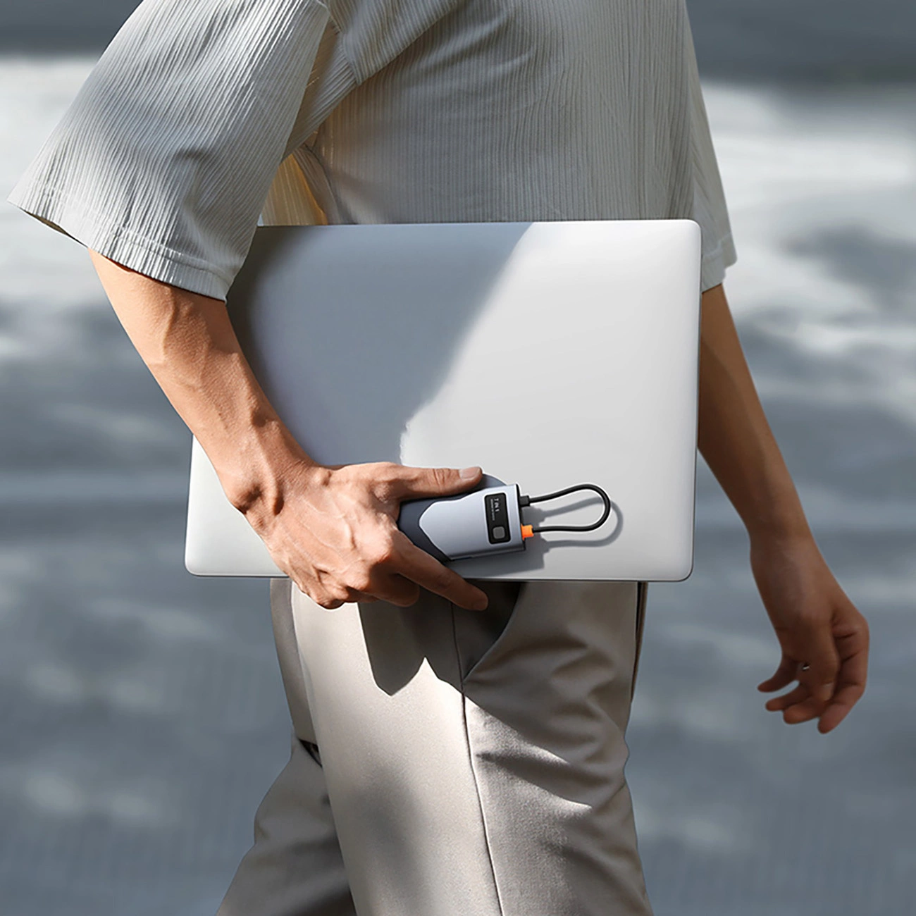 Una persona con camicia e pantaloni chiari tiene un laptop argento e un power bank all'aperto.
