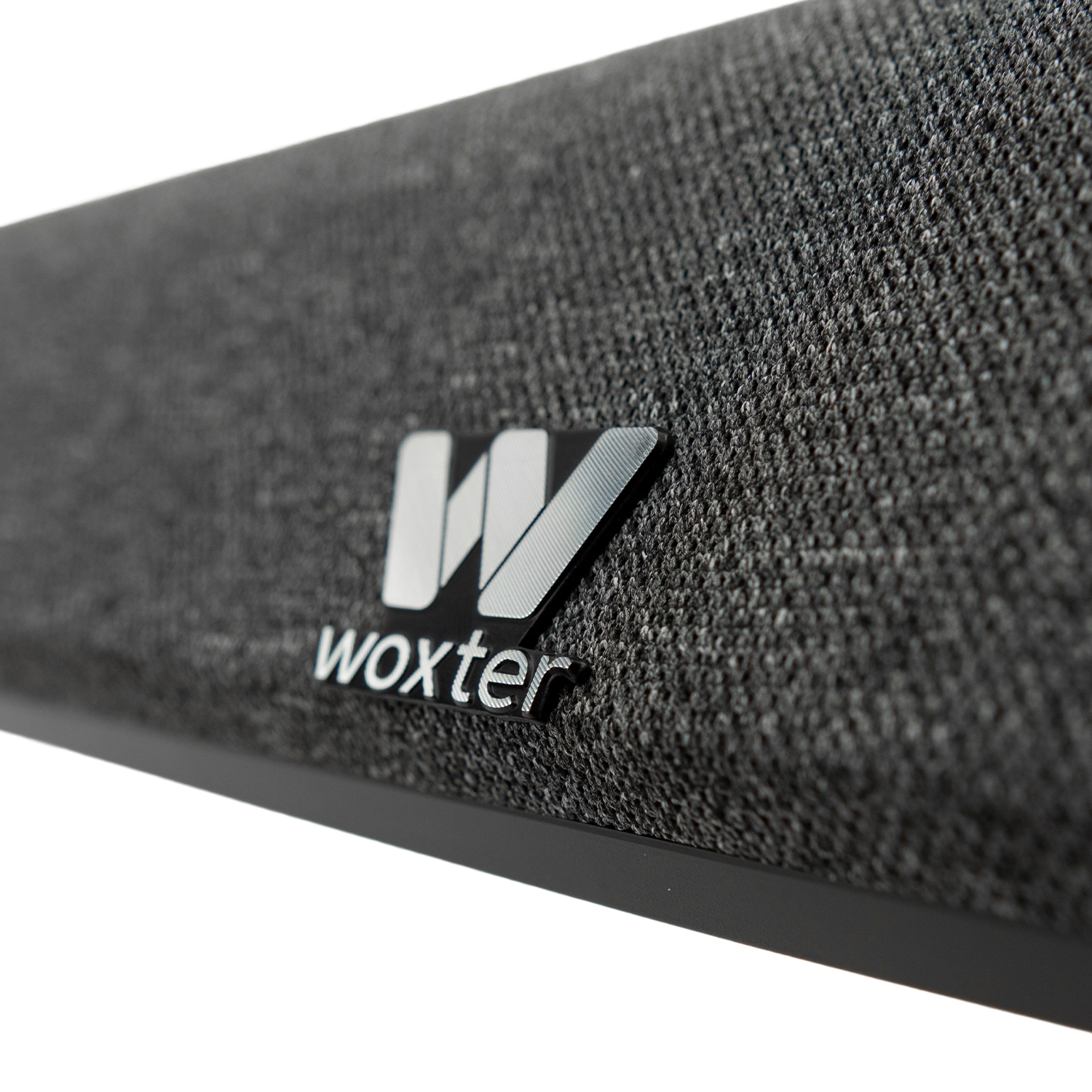 Primer plano de una barra de sonido de tela gris con el logotipo plateado de Woxter en el fondo blanco.