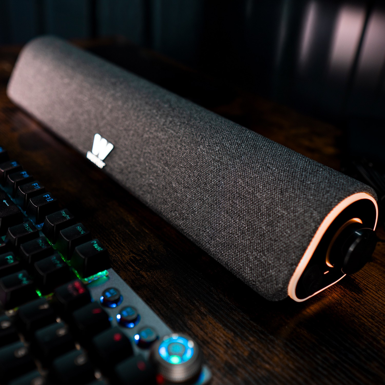 Una barra de sonido gris con borde iluminado y un teclado gaming descansan sobre un escritorio de madera.