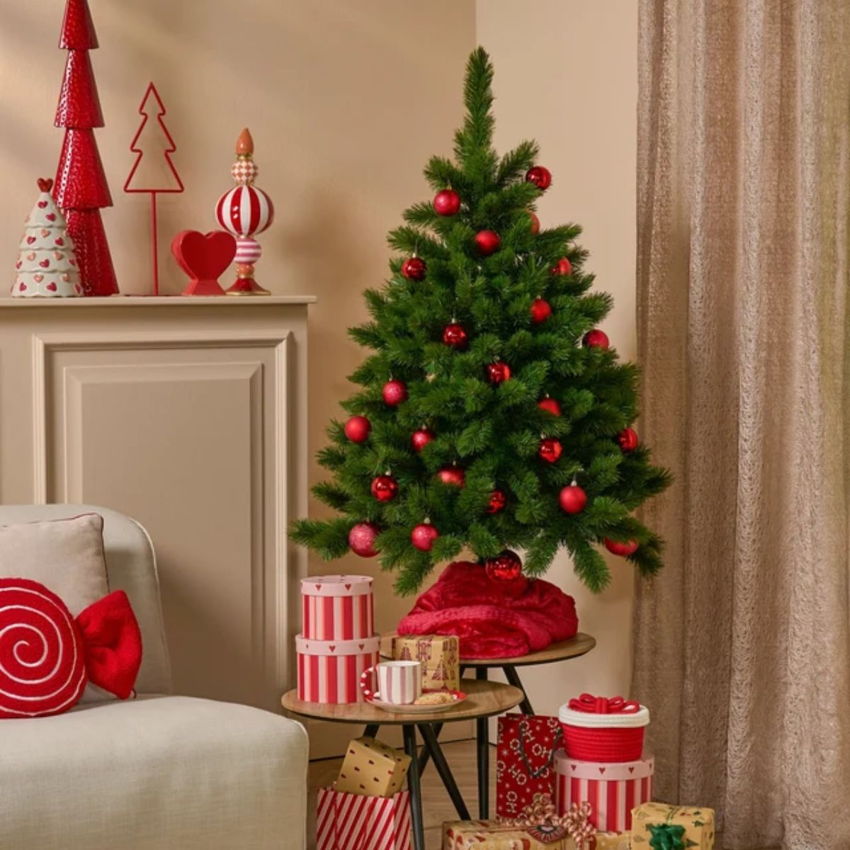 Een versierde kerstboom met rode ballen staat naast cadeautjes, een bank en rode decoratie.
