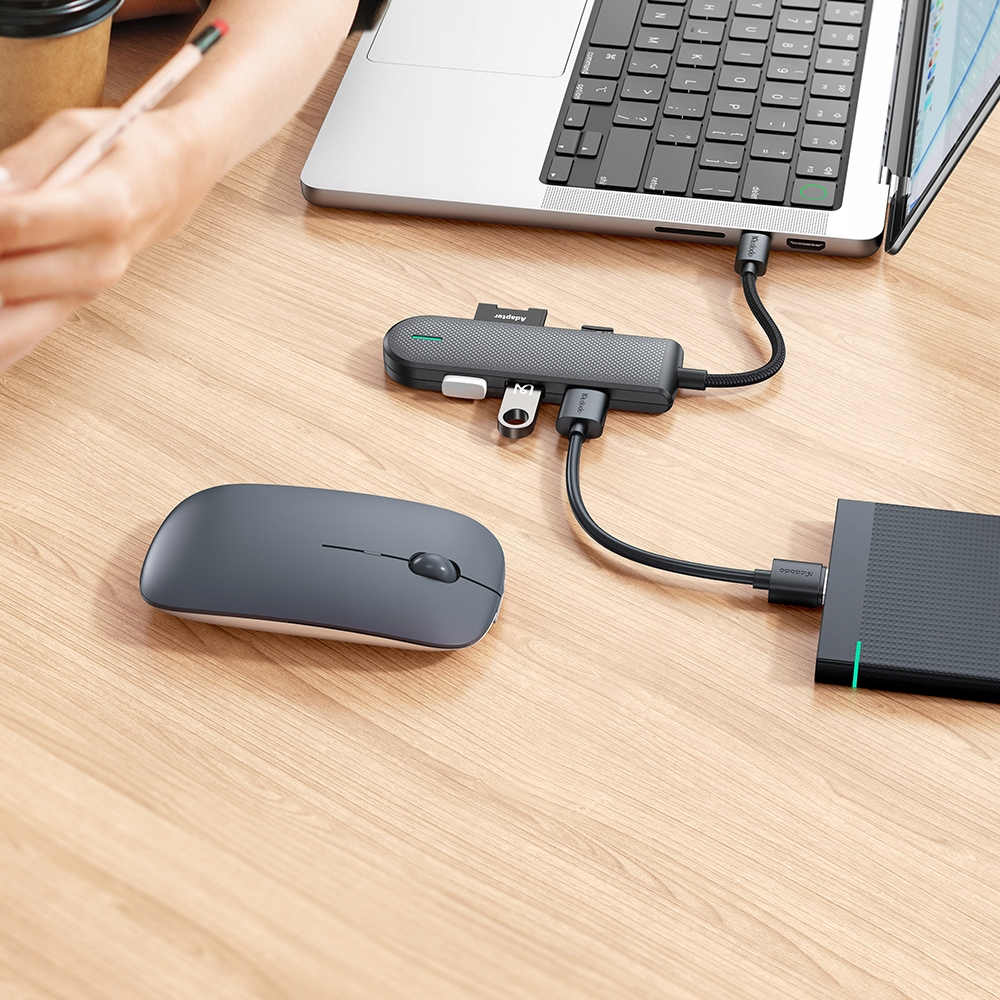 Un computer portatile, un mouse grigio, un hub USB e una memoria esterna su una scrivania di legno.