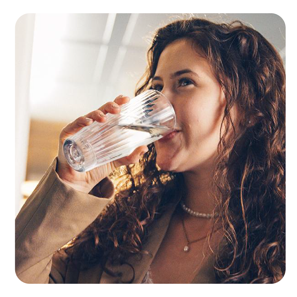 Eine Frau mit lockigem braunem Haar trinkt Wasser aus einem gerippten Glas in einem Raum mit beigem Blazer.