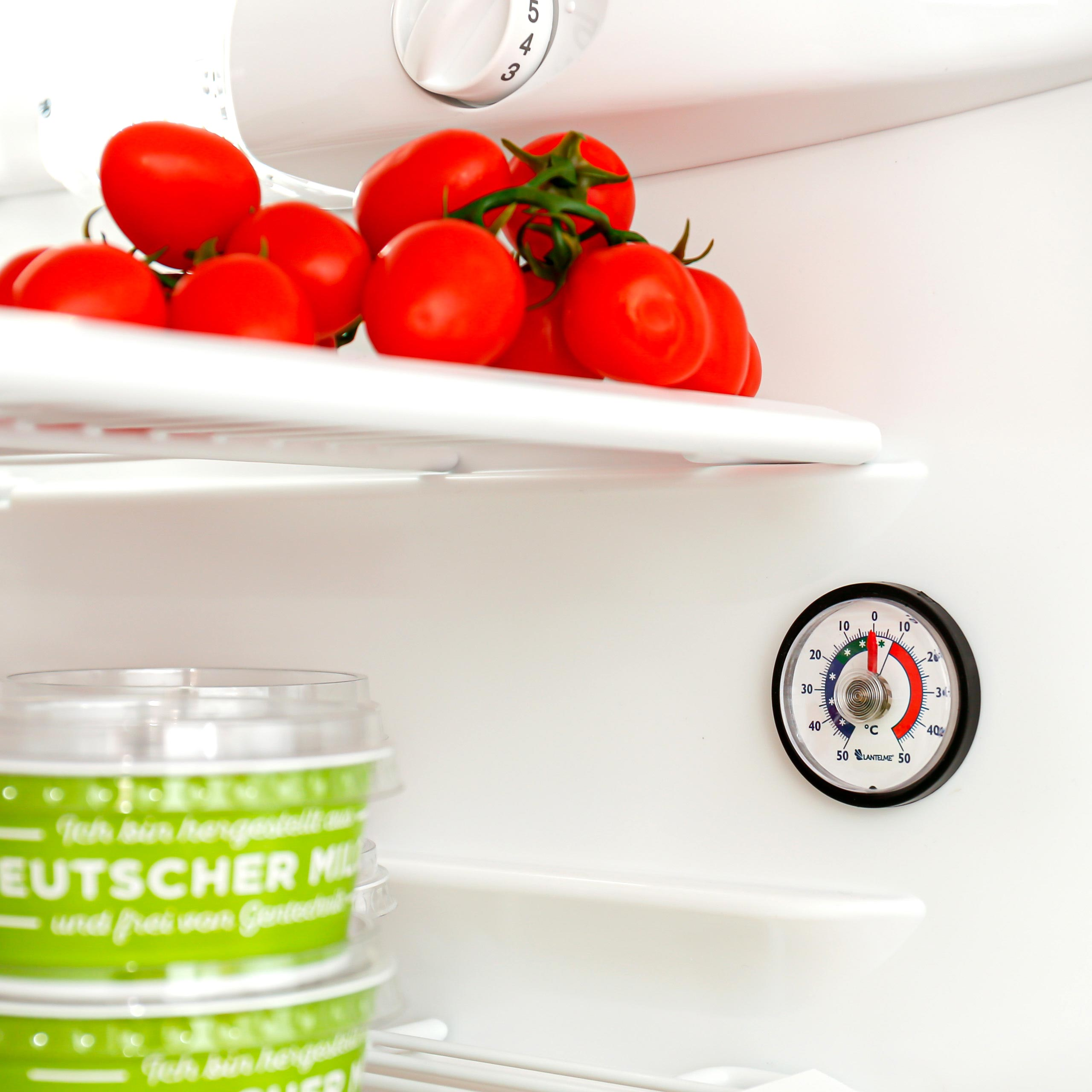 Frische Tomaten auf dem obersten und Milchprodukte auf dem unteren Regal eines weißen Kühlschranks mit Thermometer.