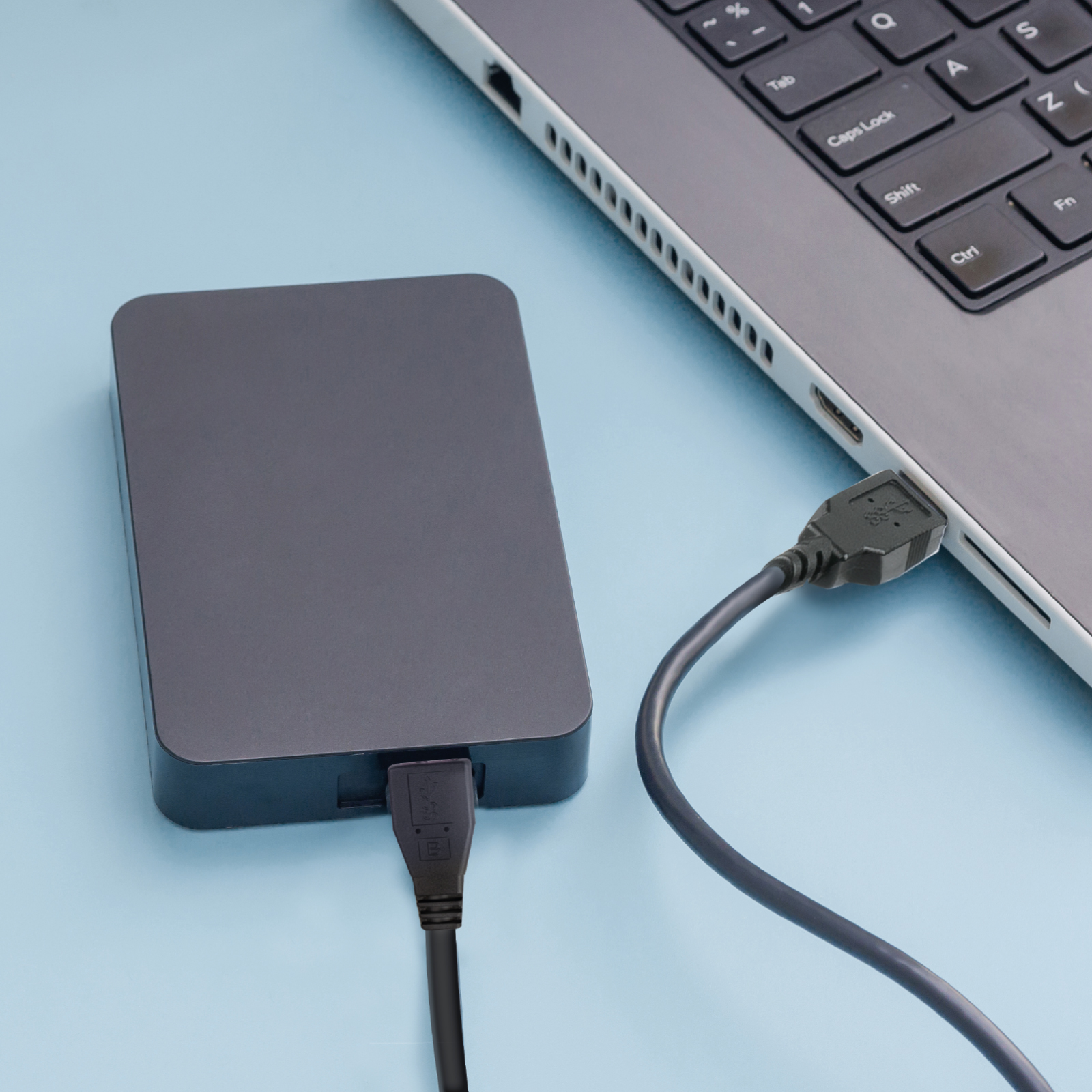 Eine graue externe Festplatte ist per USB-Kabel an einen Laptop auf hellblauem Untergrund angeschlossen.