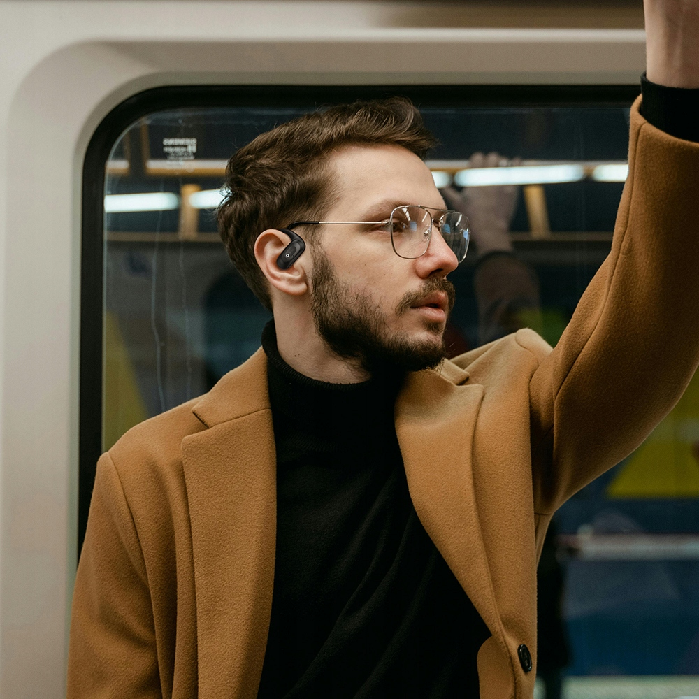 Un uomo con occhiali e auricolare nero si trova in metropolitana, indossando un cappotto marrone sopra un dolcevita nero.