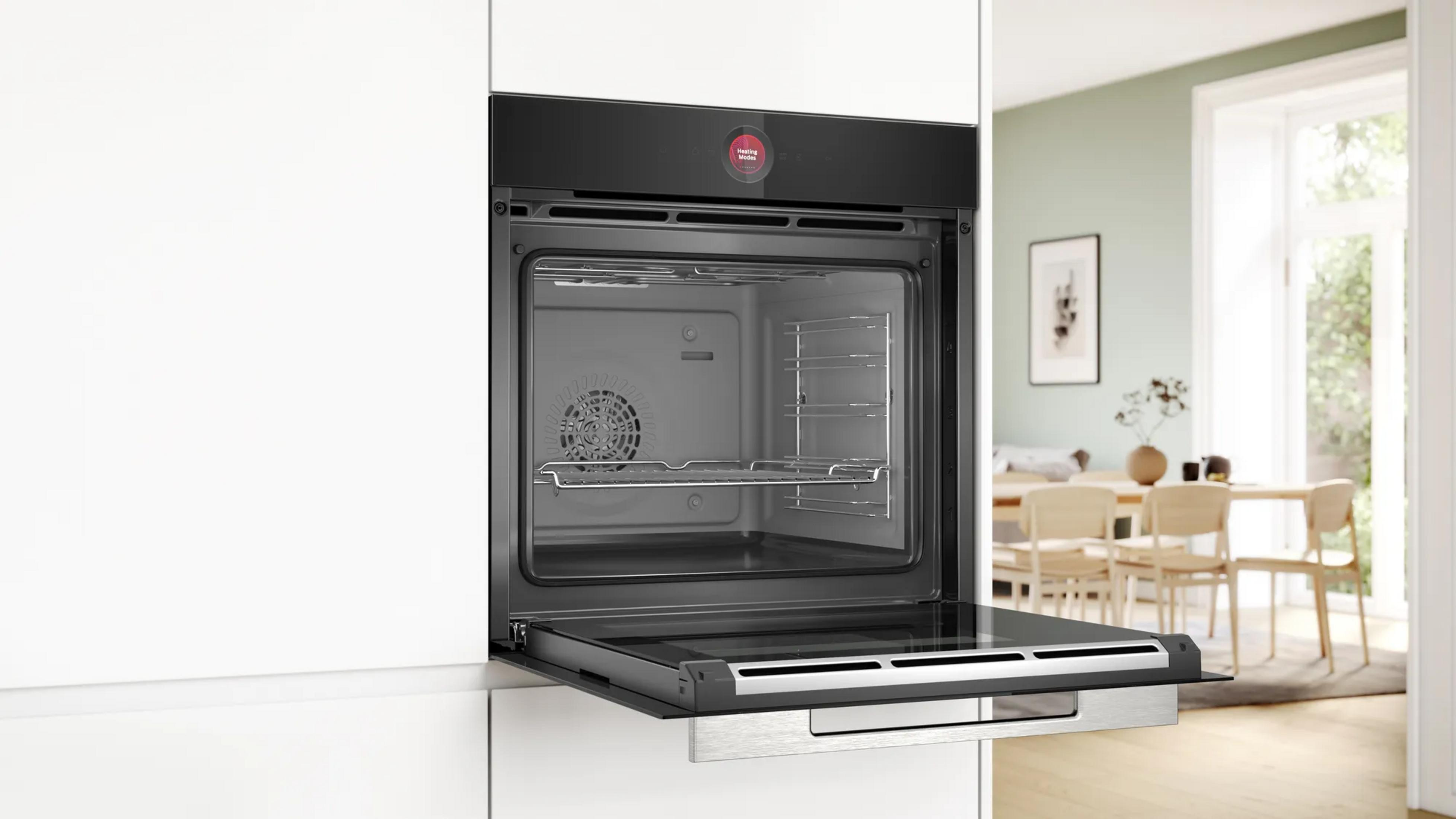 Four encastrable noir ouvert dans une cuisine blanche, avec une salle à manger lumineuse en arrière-plan.