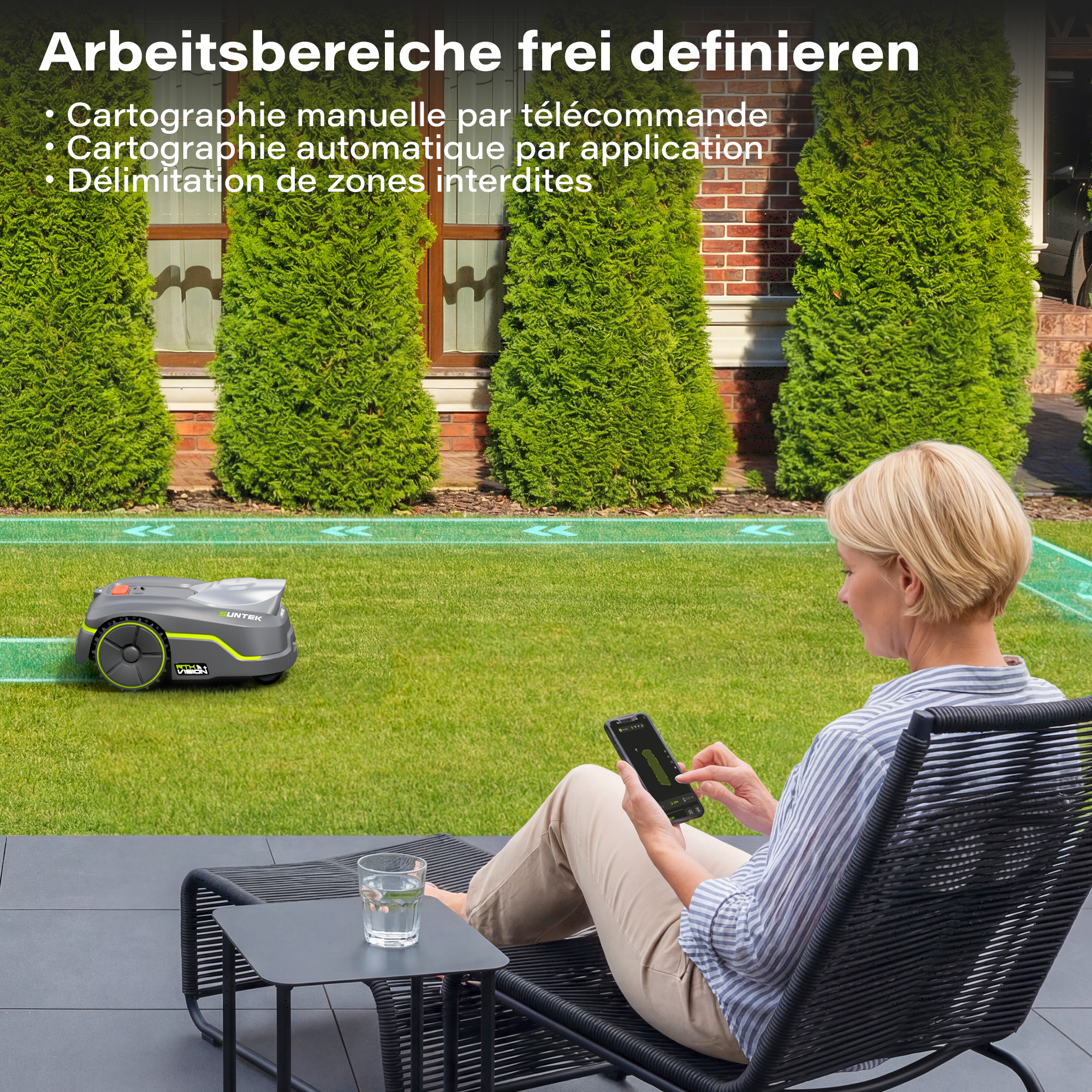 Eine Frau sitzt auf einer Liege und steuert einen Mähroboter mit einer App auf ihrem Smartphone auf dem Rasen.