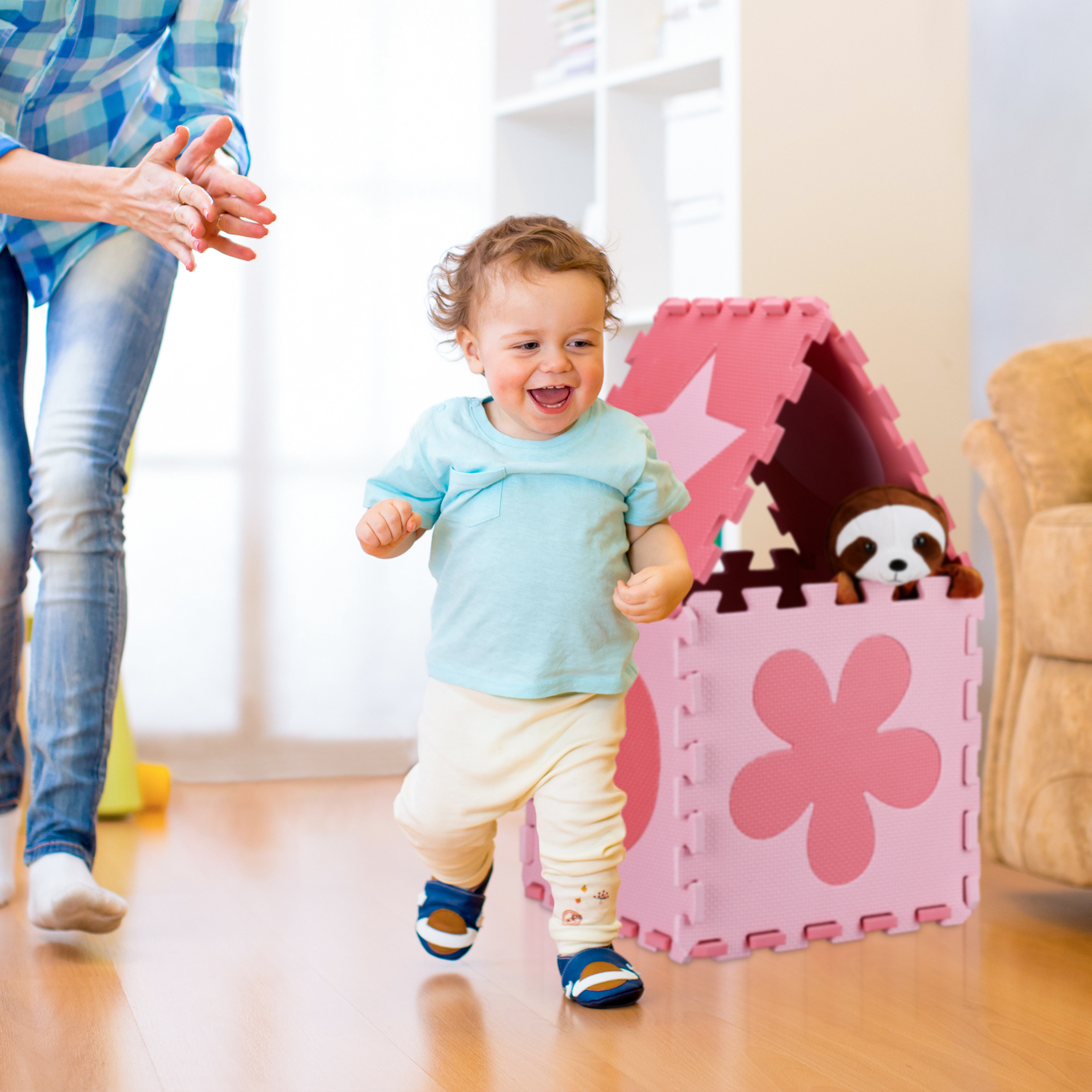 Kleinkind läuft glücklich in einem Zimmer, mit einer Mutter im Hintergrund und einem rosa Spielzeughaus.