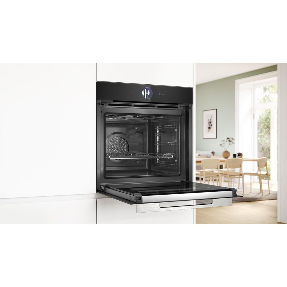Un four encastrable noir avec sa porte ouverte dans une cuisine blanche, avec une salle à manger lumineuse en arrière-plan.