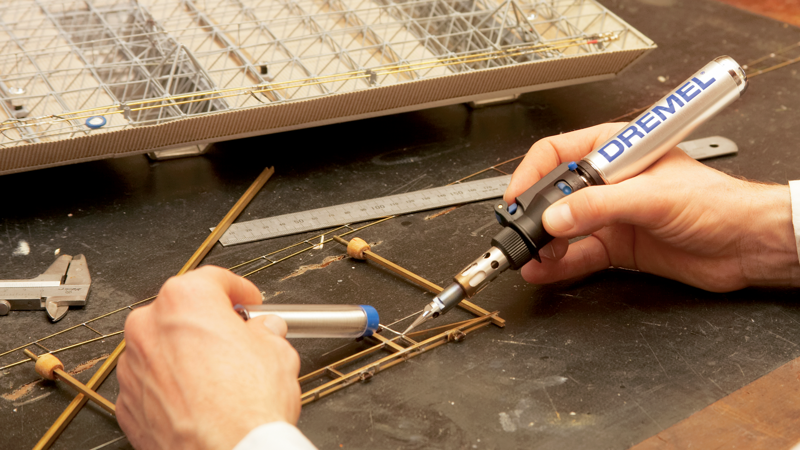 Person lötet filigrane Metallteile mit einem Dremel-Lötkolben an einem Werktisch.