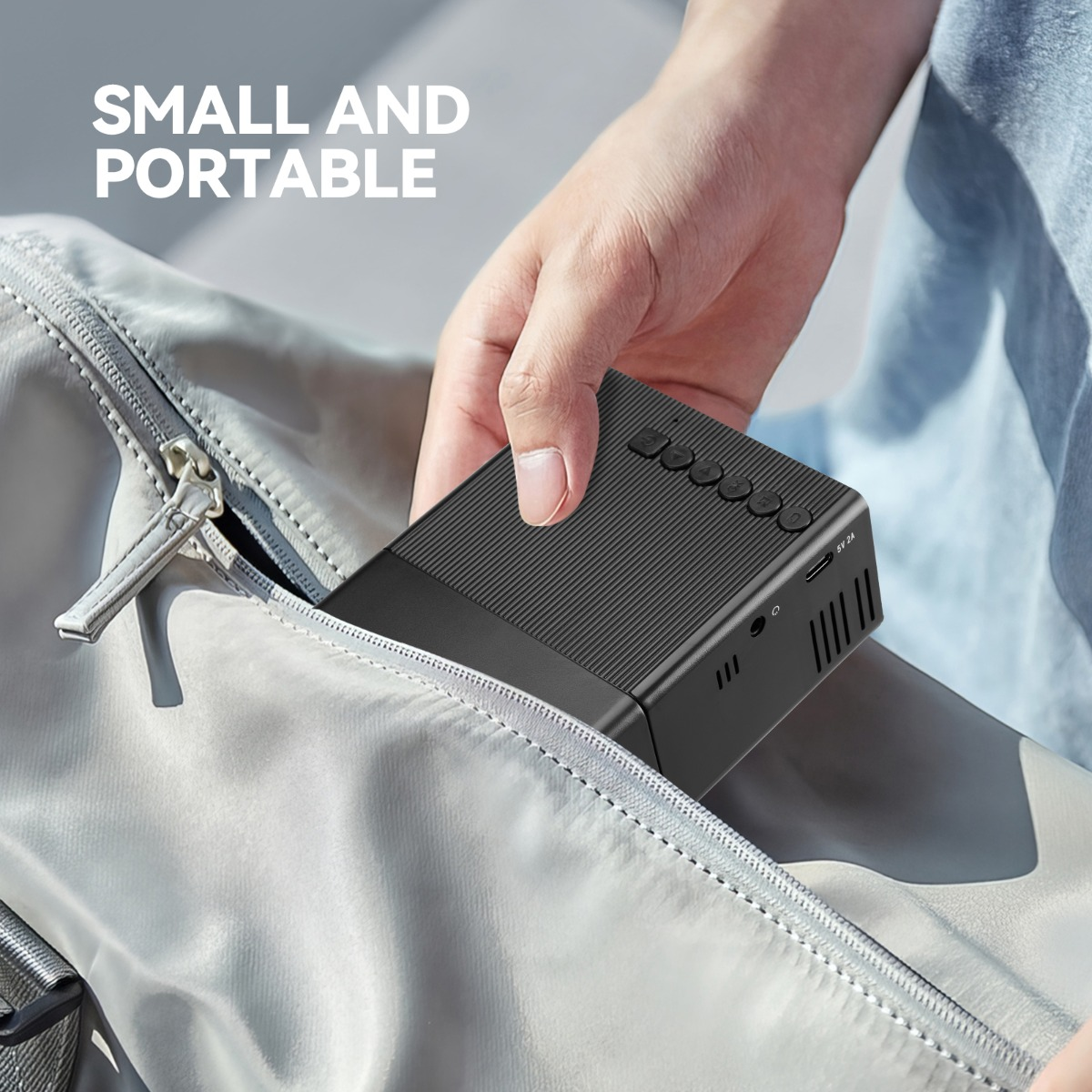 Eine Hand legt einen kleinen, schwarzen Bluetooth-Lautsprecher in eine helle, offene Tasche.