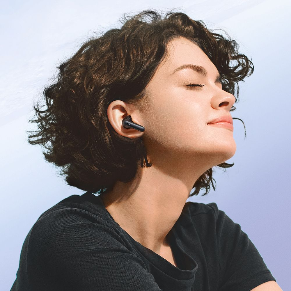 Una donna con capelli corti e ricci indossa auricolari wireless neri su uno sfondo azzurro.